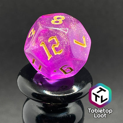 A close up of the D12 from the Moxie 7 piece dice set from Tabletop Loot; fuchsia dice packed with glitter and gold gothic numbering.