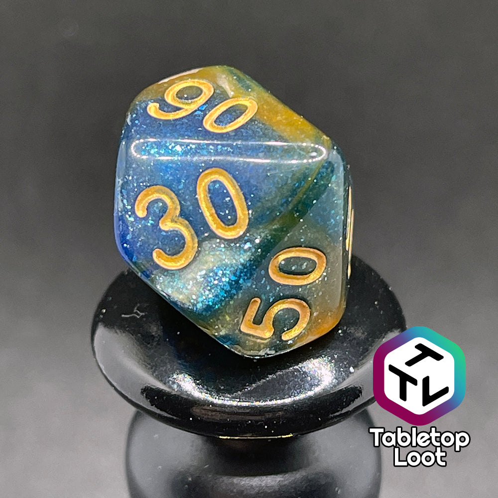 A close up of the percentile die from the Nimrodel 7 piece dice set from Tabletop Loot with swirls of glittering blue, green, and orange and golden numbering.