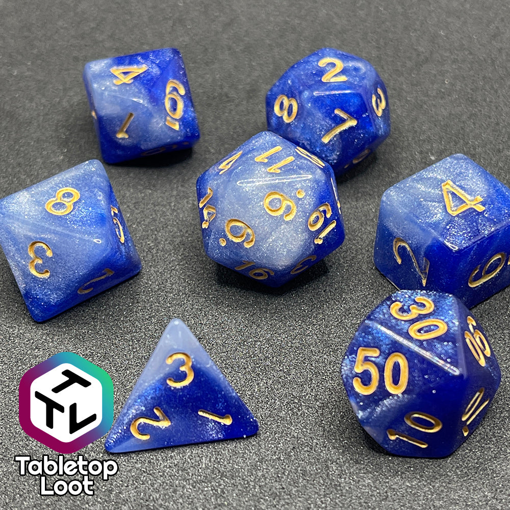 A close up of the Queen's Treasure 7 piece dice set from Tabletop Loot packed with glitter in swirls of white and royal blue and gold numbering.