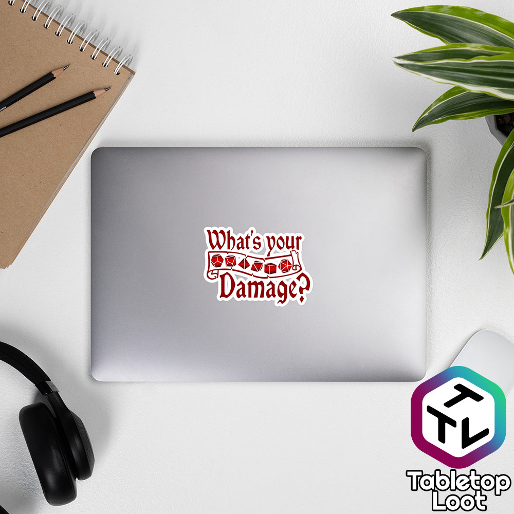 A 4 inch sticker from Tabletop Loot on a laptop says "What's your Damage?" in an Old English script around a banner of polyhedral dice in red and black.