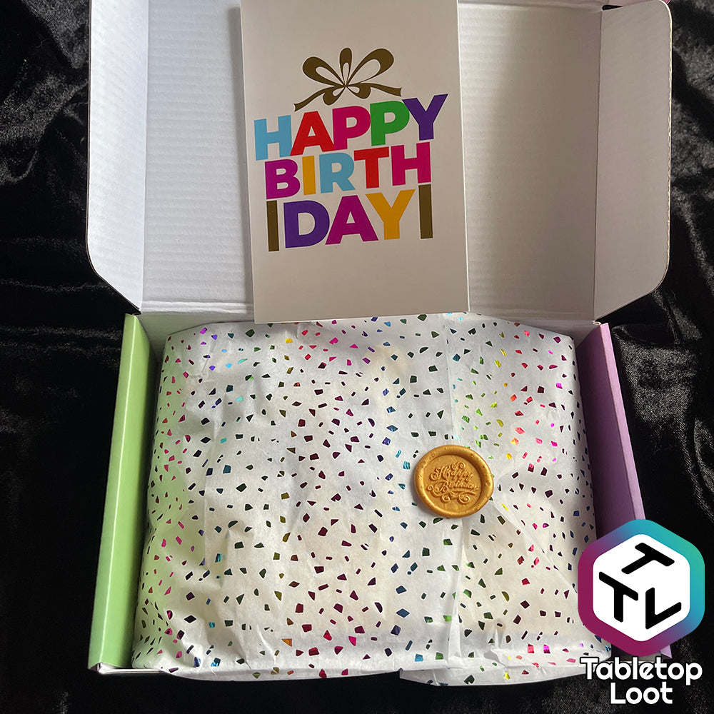 Color tissue paper sits in a box and is held closed by a gold wax seal that reads "Happy Birthday". Above the paper, leaning on the box's open lid is a birthday card.
