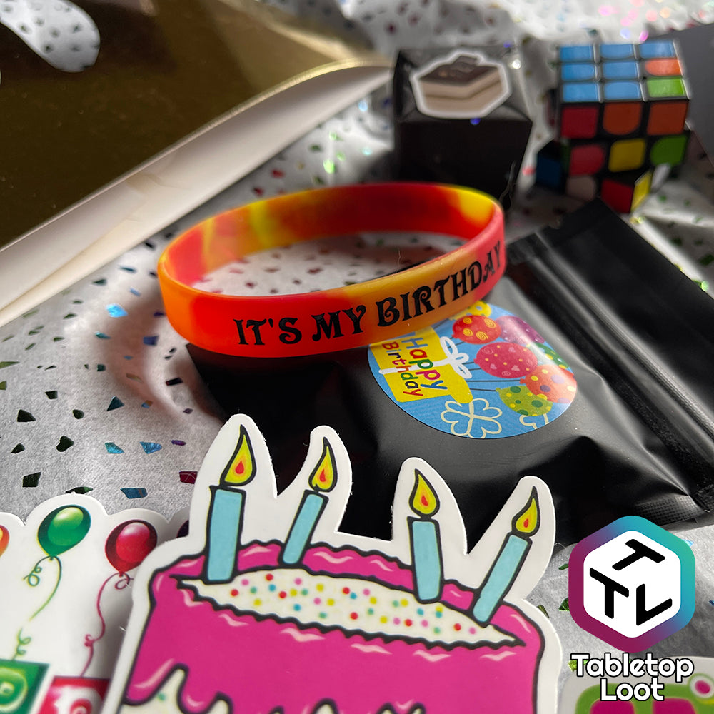 An orange and red silicone bracelet with the words "It's my birthday" written in black letters sits on top of a black bag with a birthday sticker on it.