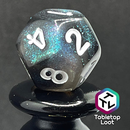 A close up of the D12 from the Black Moonstone 7 piece dice set from Tabletop Loot with swirls of black and teal iridescent glitter, inked in white.