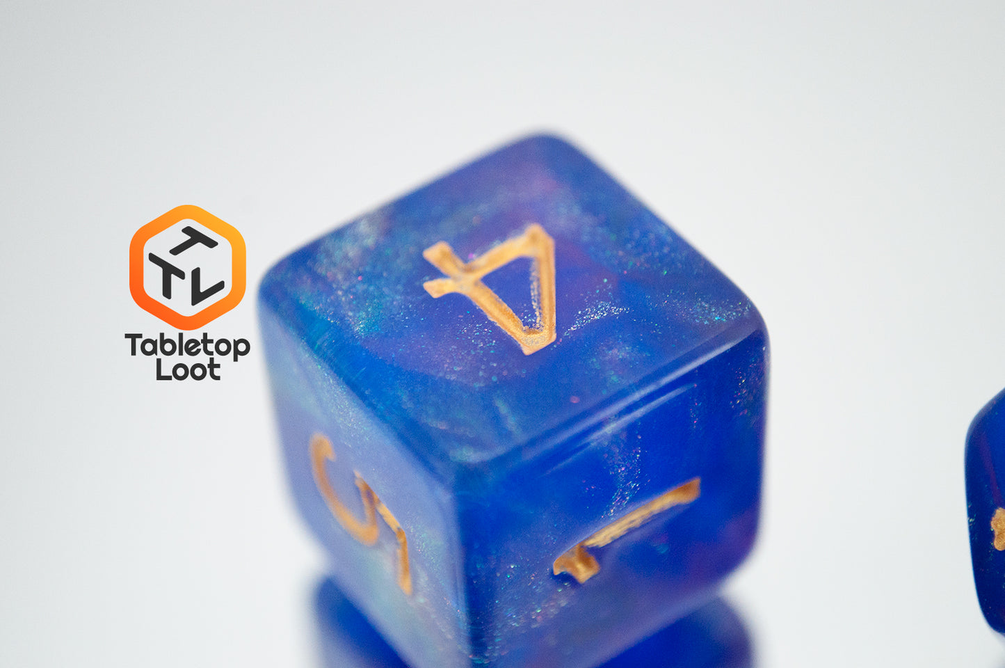 A close up of the D6 from the Blue Enchantress 7 piece dice set from Tabletop Loot with shimmering shades of pink, purple, and blue swirling resin and gold numbering.