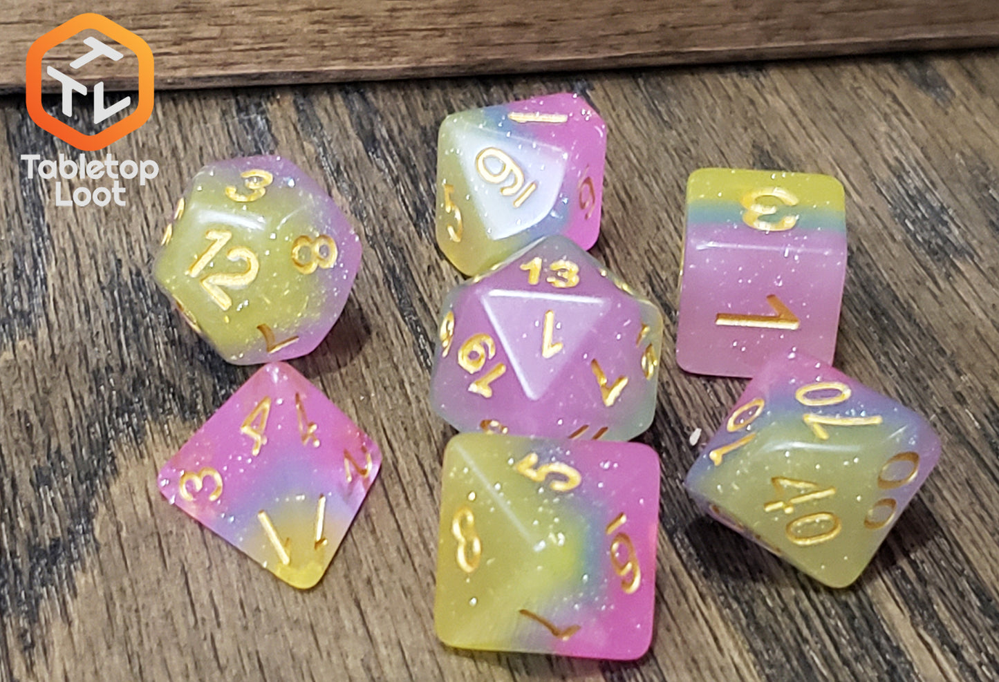 The Candyland 7 piece dice set from Tabletop Loot with layers of yellow, blue, and pink glittery resin and gold numbering.