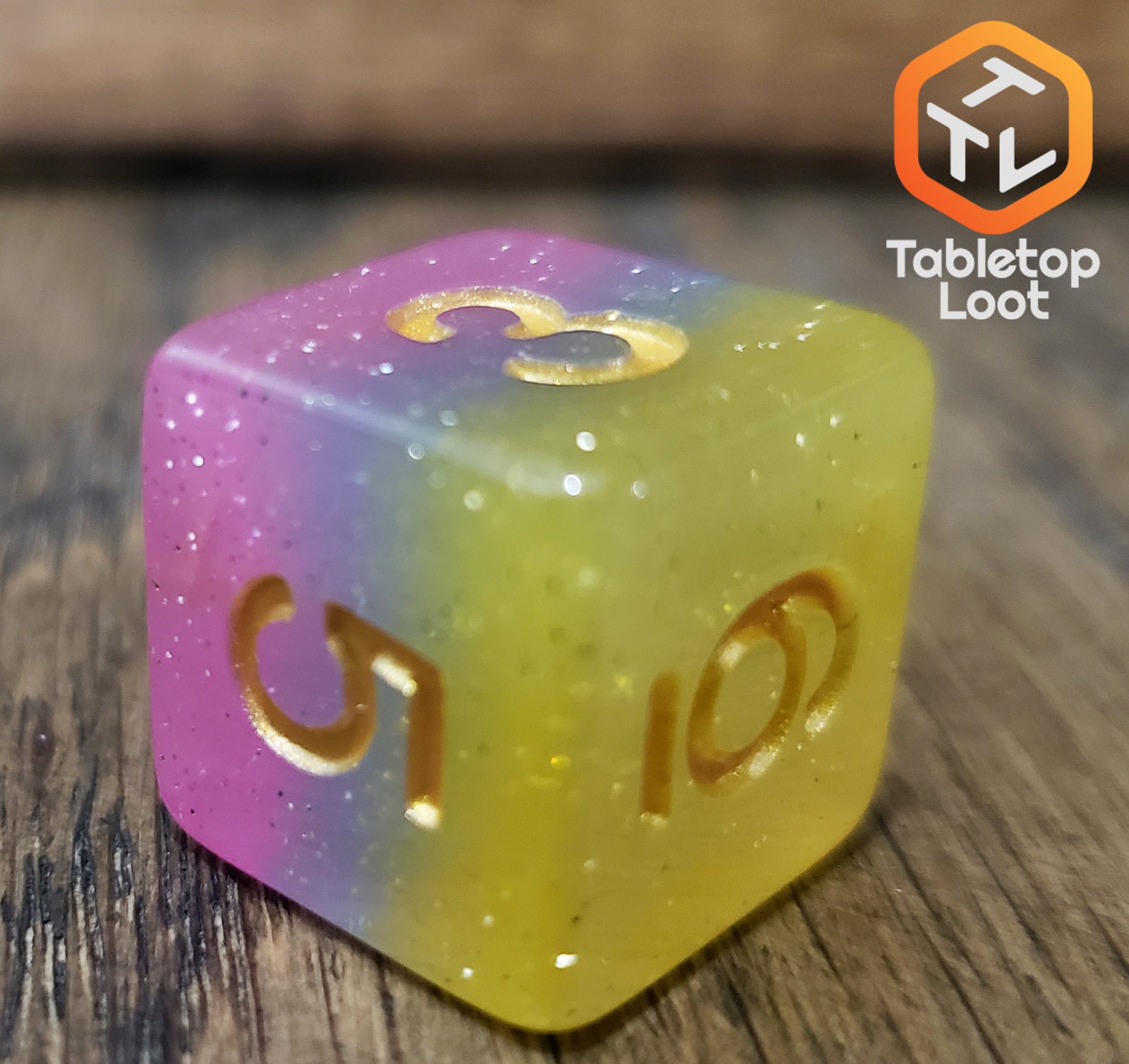 A close up of the D6 from the Candyland 7 piece dice set from Tabletop Loot with layers of yellow, blue, and pink glittery resin and gold numbering.