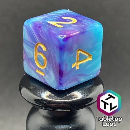 A close up of the D6 from the Cotton Candy 7 piece dice set from Tabletop Loot with swirls of blue and purple and golden numbers.
