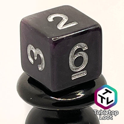 A close up of the D6 from the Darkness 7 piece dice set from Tabletop Loot; translucent purple so dark it looks black except where the light peeks through in corners, inked in silver.