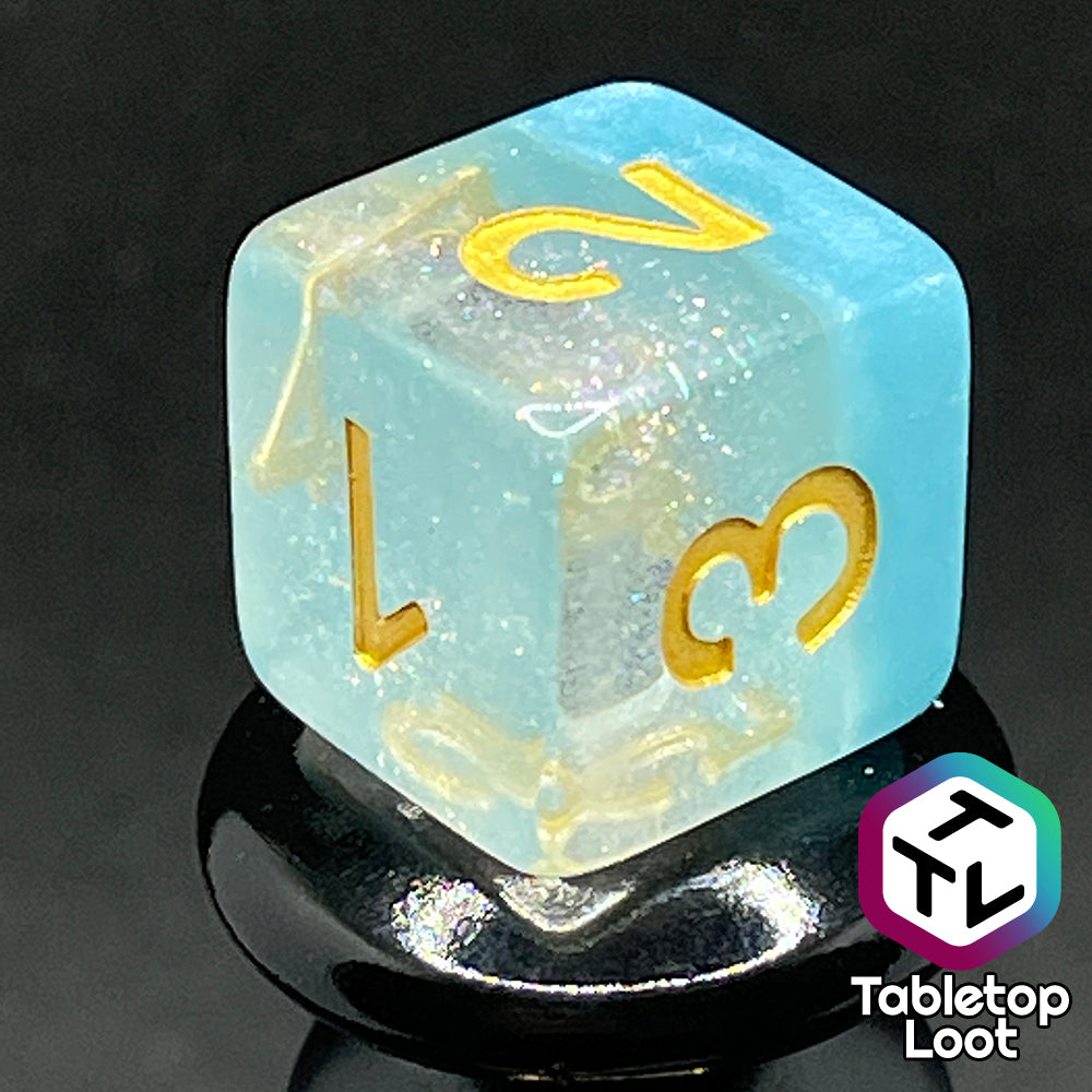 A close up of the D6 from the Elsa 7 piece dice set from Tabletop Loot with a layer of sky blue under glittery clear resin and golden numbering.