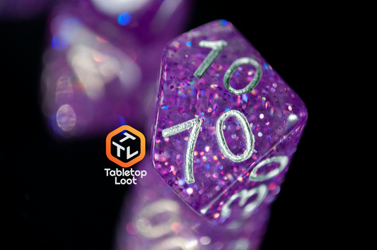 A close up of the percentile die from the Faerie Fire 7 piece dice set from Tabletop Loot packed with purple and blue iridescent sparkles in translucent purple with silver numbering.
