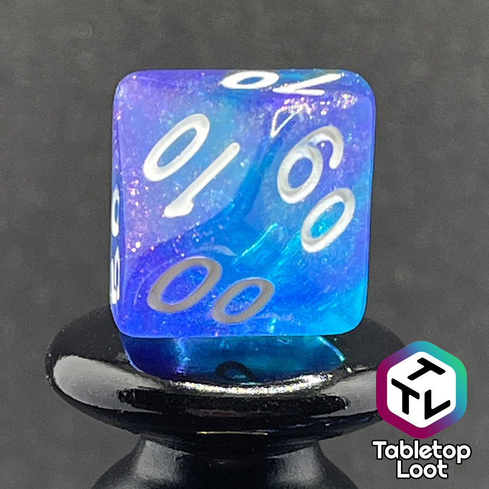 A close up of the percentile die from the Fathomless 7 piece dice set from Tabletop Loot with swirls of glitter and purple in deep blue and white numbering.