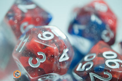 A close up of the Fire and Ice 7 piece dice set from Tabletop Loot with swirls of blue and red in a clear glittery resin and silver numbering.