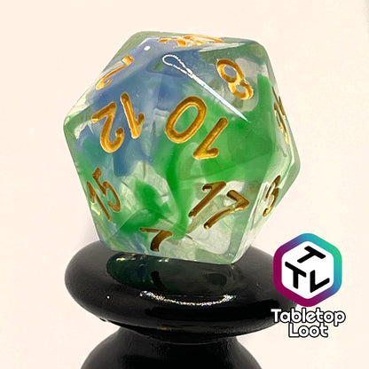 A close up of the D20 from the Gentle Repose 7 piece dice set with swirls of bright green and blue in clear resin and gold numbering.