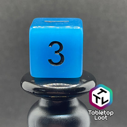 A close up of the D6 from the Ghost Ice 7 piece dice set from Tabletop Loot with glowing blue pigment and black numbers, shown in the light.