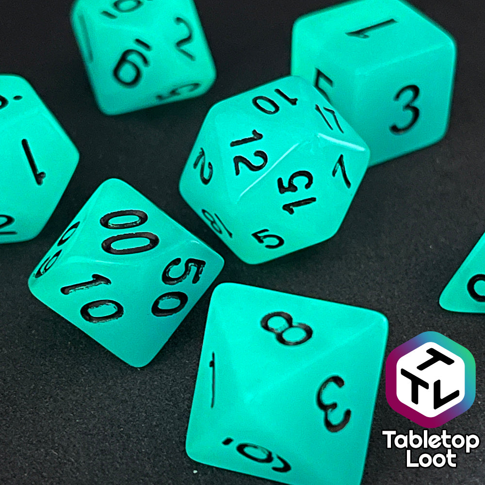 A close up of the Ghost Ice 7 piece dice set from Tabletop Loot with glowing blue pigment and black numbers, shown in the dark.