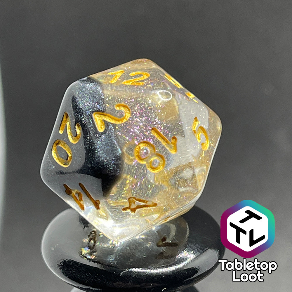 A close up of the D20 from the Inkwell 7 piece dice set from Tabletop Loot with a layer of black glittery color on one side of the clear glittery resin and gold numbering.
