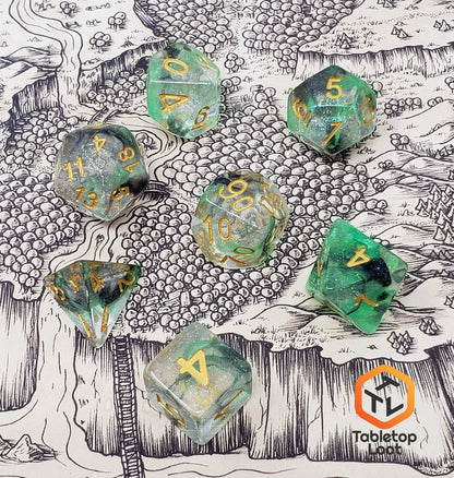 The Luminous Venom 7 piece dice set from Tabletop Loot with green and black swirls in a clear glittery resin with gold numbering on a map by Deven Rue.