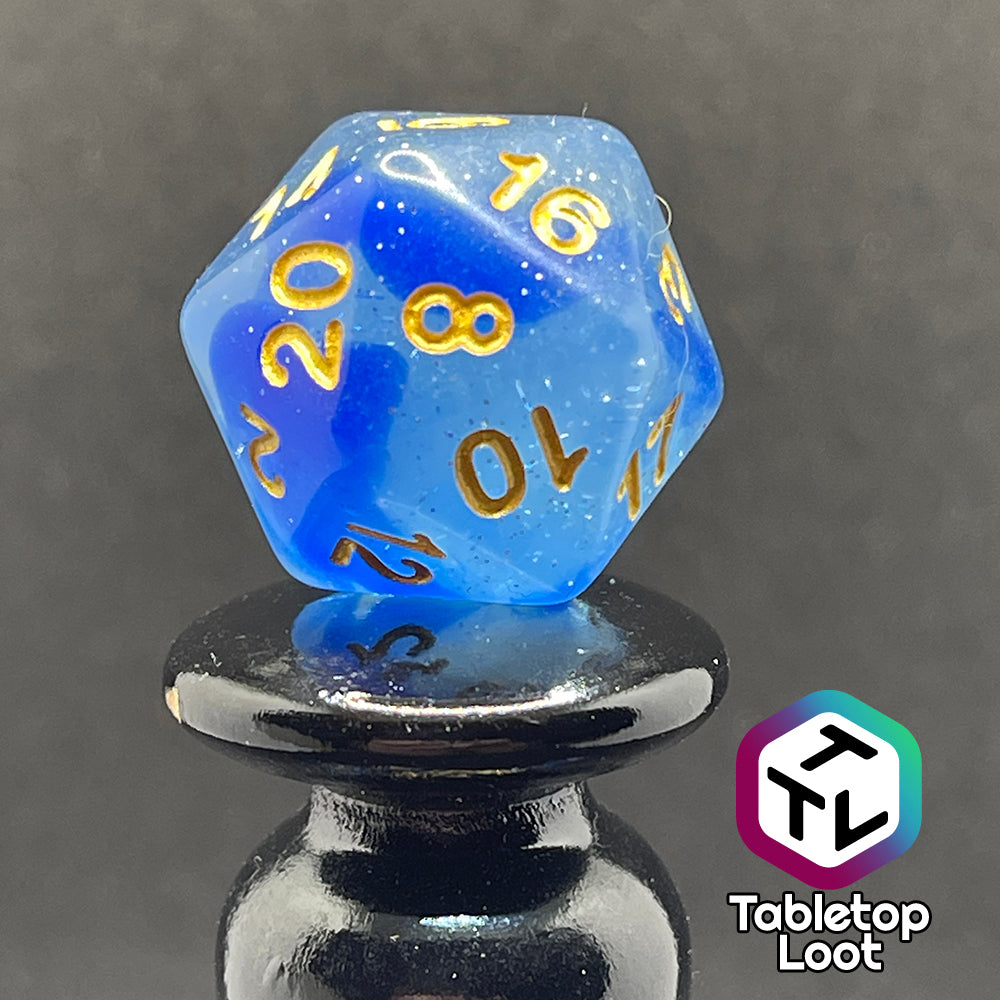 A close up of the D20 from the Mermaid's Crown 7 piece dice set from Tabletop Loot with swirled blue tones, tons of glitter, and gold numbering.