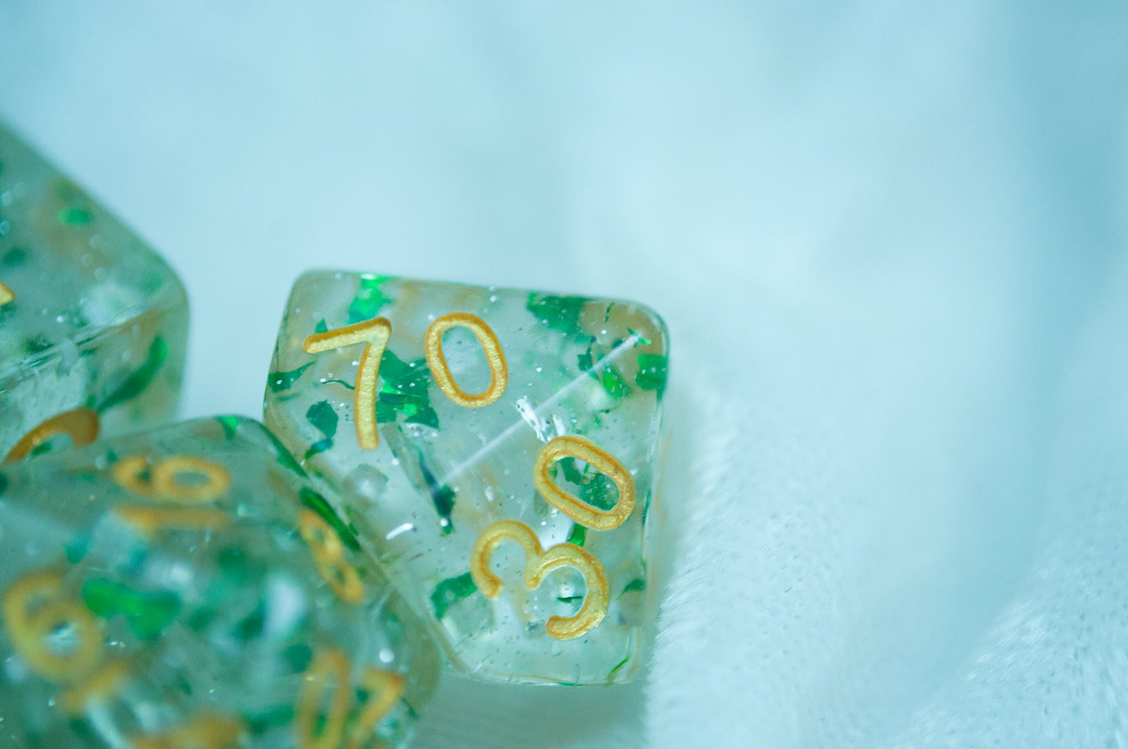 A close up of the Metallic Emerald 7 piece dice set from Tabletop Loot with green metallic flakes suspended in clear resin with glitter and gold numbering.