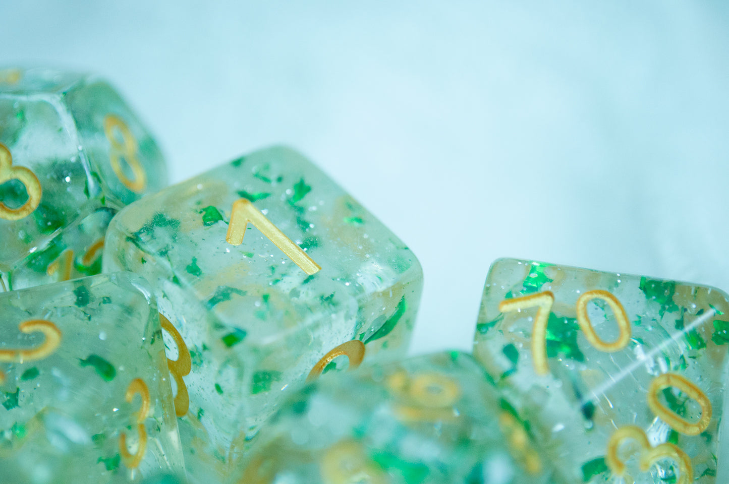 A close up of the Metallic Emerald 7 piece dice set from Tabletop Loot with green metallic flakes suspended in clear resin with glitter and gold numbering.