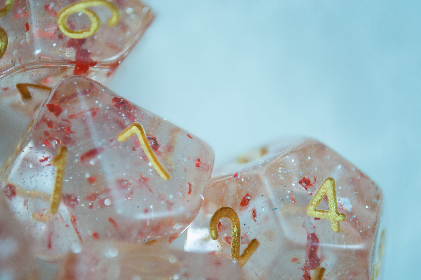 A close up of the Metallic Ruby 7 piece dice set from Tabletop Loot with red metallic flakes suspended in clear resin with glitter and gold numbering.