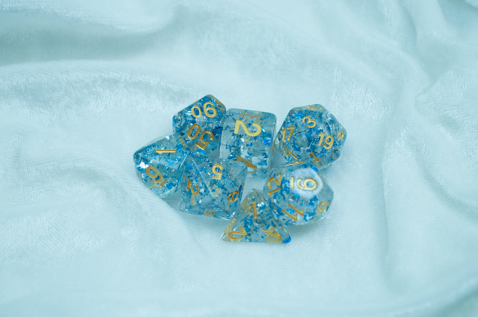 The Metallic Sapphire 7 piece dice set from Tabletop Loot with blue metallic flakes suspended in clear resin with glitter and gold numbering.