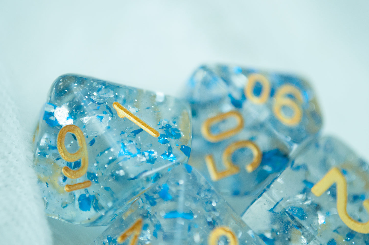 A close up of the Metallic Sapphire 7 piece dice set from Tabletop Loot with blue metallic flakes suspended in clear resin with glitter and gold numbering.