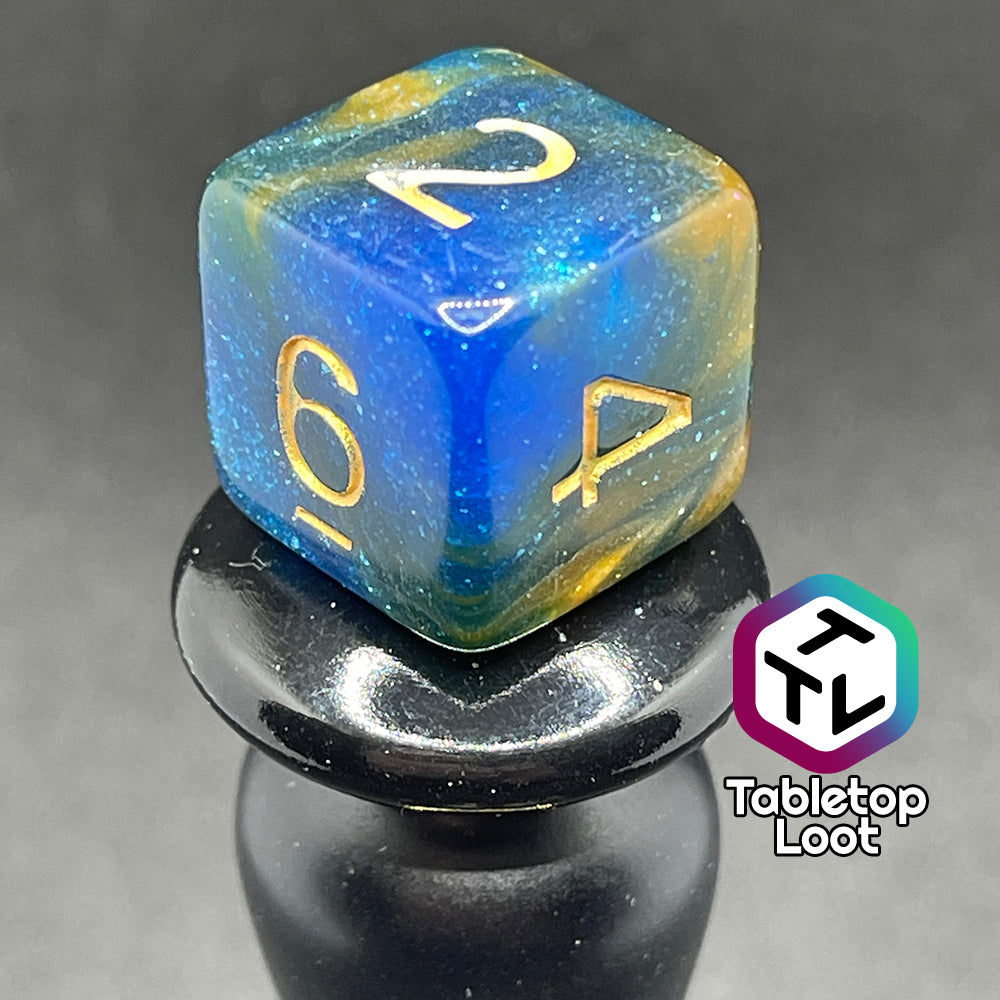 A close up of the D6 from the Nimrodel 7 piece dice set from Tabletop Loot with swirls of glittering blue, green, and orange and golden numbering.
