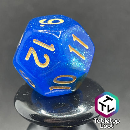 A close up of the D12 from the Nimrodel 7 piece dice set from Tabletop Loot with swirls of glittering blue, green, and orange and golden numbering.