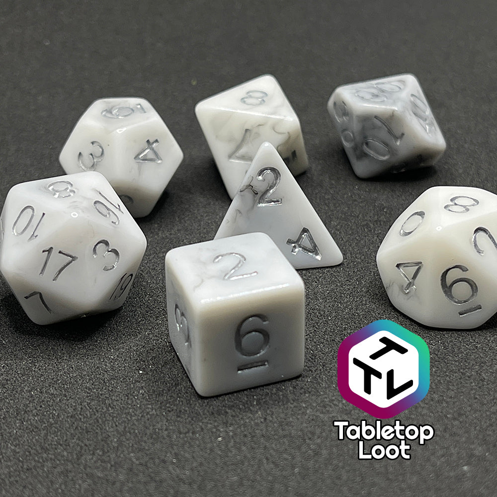 A close up of the Oath Breaker 7 piece dice set from Tabletop Loot; opaque white with swirls of silver peeking out and silver numbering.