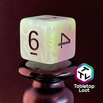 A close up of the D6 from the Retro 80s glow in the dark 7 piece dice set from Tabletop Loot with swirls of pink and yellow glow pigment and black numbering, shown glowing.