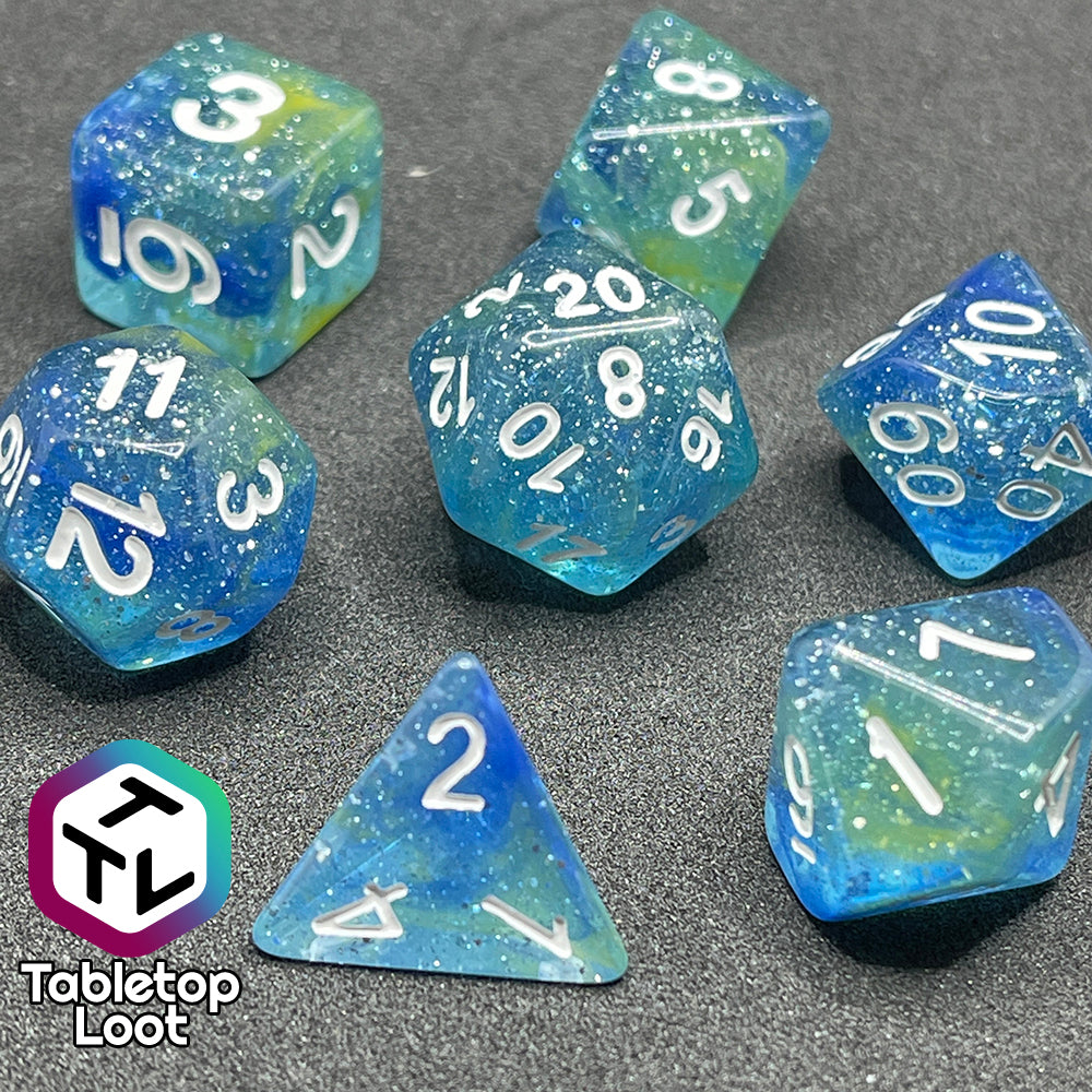 A close up of the Stellar Dust 7 piece dice set from Tabletop Loot with wisps of blue and yellow in clear resin and large sparkles with white numbering.