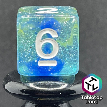 A close up of the D6 from the Stellar Dust 7 piece dice set from Tabletop Loot with wisps of blue and yellow in clear resin and large sparkles with white numbering.