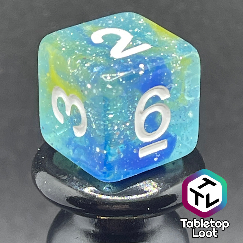 A close up of the D6 from the Stellar Dust 7 piece dice set from Tabletop Loot with wisps of blue and yellow in clear resin and large sparkles with white numbering.