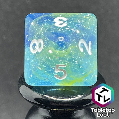 A close up of the D8 from the Stellar Dust 7 piece dice set from Tabletop Loot with wisps of blue and yellow in clear resin and large sparkles with white numbering.