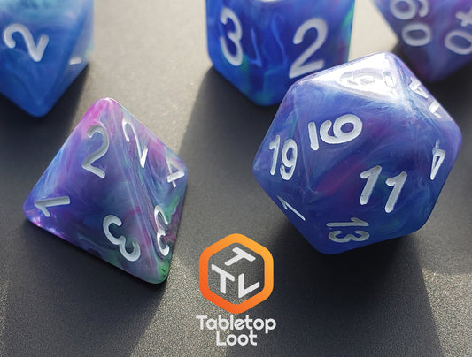 A close up of the Dancing Lights 7 piece dice set from Tabletop Loot with swirls of purple, pink, green, teal, and blue resin and white numbers.