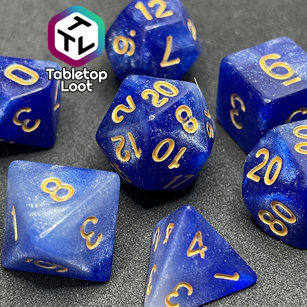 A close up of the Queen's Treasure 7 piece dice set from Tabletop Loot packed with glitter in swirls of white and royal blue and gold numbering.