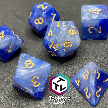 A close up of the Queen's Treasure 7 piece dice set from Tabletop Loot packed with glitter in swirls of white and royal blue and gold numbering.