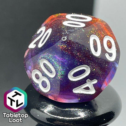A close up of the percentile die from the Transmutation 7 piece dice set from Tabletop Loot with swirls of blue and red appearing purple in some spots, filled with iridescent glitter adding little rainbows in places, and inked in white.