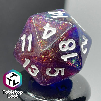 A close up of the D20 from the Transmutation 7 piece dice set from Tabletop Loot with swirls of blue and red appearing purple in some spots, filled with iridescent glitter adding little rainbows in places, and inked in white.